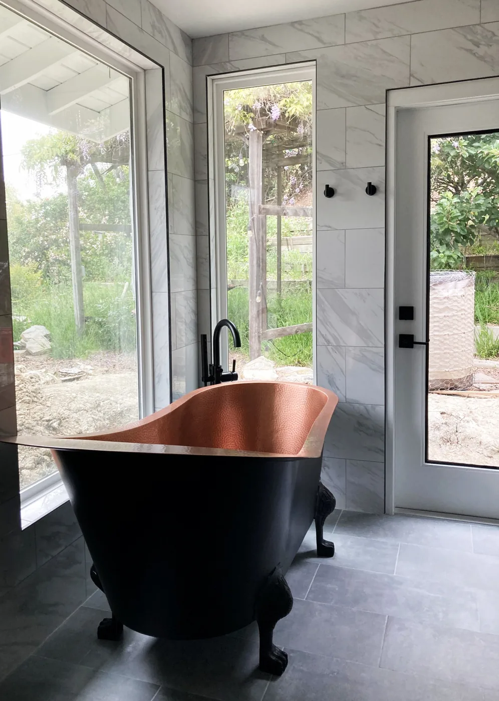 Copper-interior clawfoot tub with black exterior against marble wall tile and large windows
