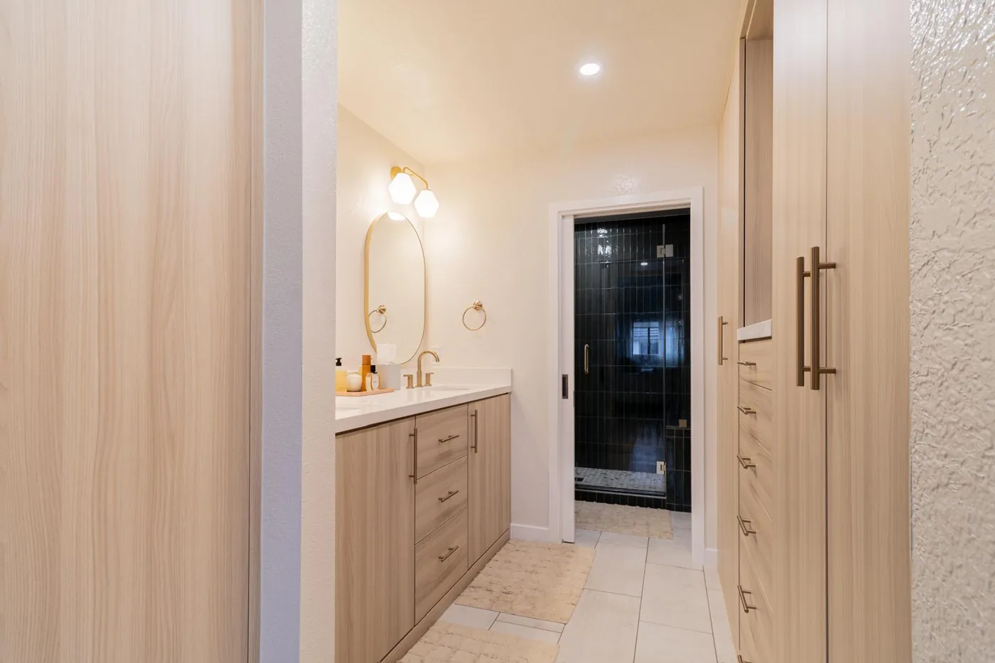 Bathroom with light wood vanity, oval mirror, white countertop and dark tile shower beyond