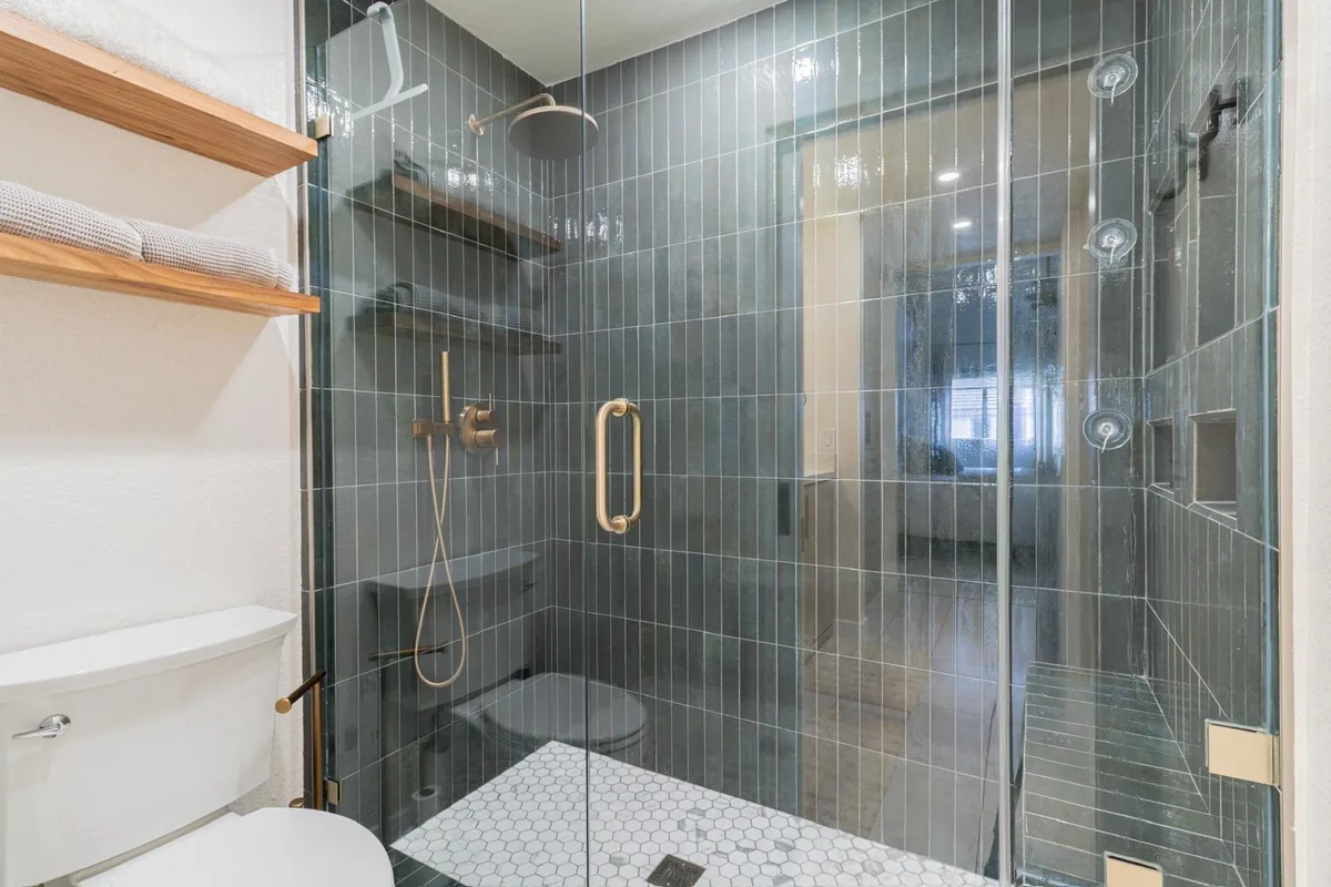 Walk-in shower with green vertical tile, brass fixtures, frameless glass and hexagon marble floor
